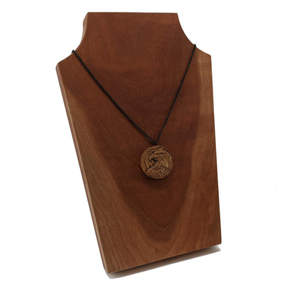 Round wooden necklace pendant carved in the shape of an eagles head. Made from hard maple wood. Hanging from a black stainless steel chain against a cherry wood background.