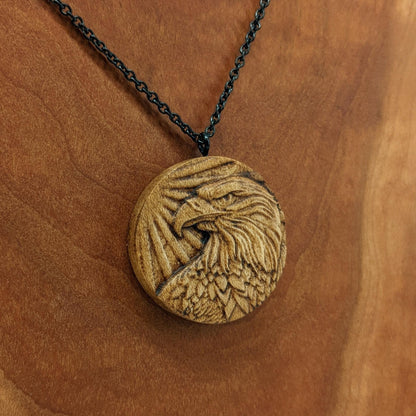 Round wooden necklace pendant carved in the shape of an eagles head. Made from hard maple wood. Hanging from a black stainless steel chain against a cherry wood background.