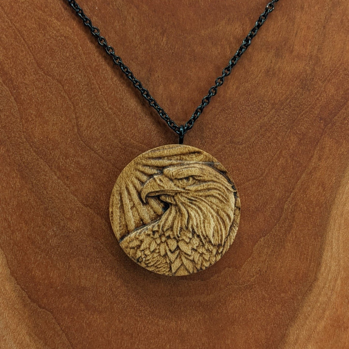 Round wooden necklace pendant carved in the shape of an eagles head. Made from hard maple wood. Hanging from a black stainless steel chain against a cherry wood background.