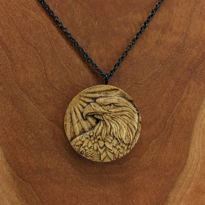 Round wooden necklace pendant carved in the shape of an eagles head. Made from hard maple wood. Hanging from a black stainless steel chain against a cherry wood background.