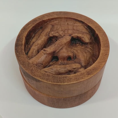 Round wooden box 3D carved with a recessed carving of an old mans face with many wrinkles and large eyebrows, with his fingers spread across and covering the right side of his face. Made from hard maple wood stained brown against a white background rotating on a table.