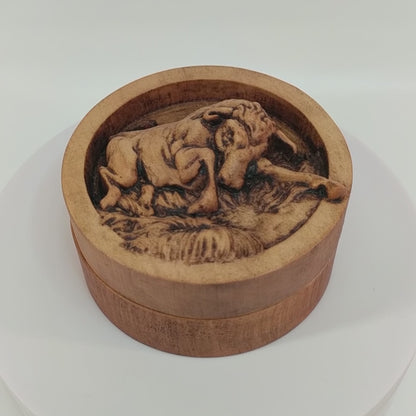 Round wooden box 3D carved with a angry bull charging forward stomping over small grassy patches on the ground. Its head is angled downward with its curved horns pointed ahead. Made from hard maple wood stained brown against a white background rotating on a table.