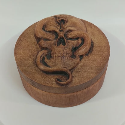 Round wooden box 3D carved with a Large skull facing forward with a snake intertwined through its jaw bone and eye sockets. The snake is looking forward hissing with its tongue out. Made from hard maple wood stained brown against a white background rotating on a table.