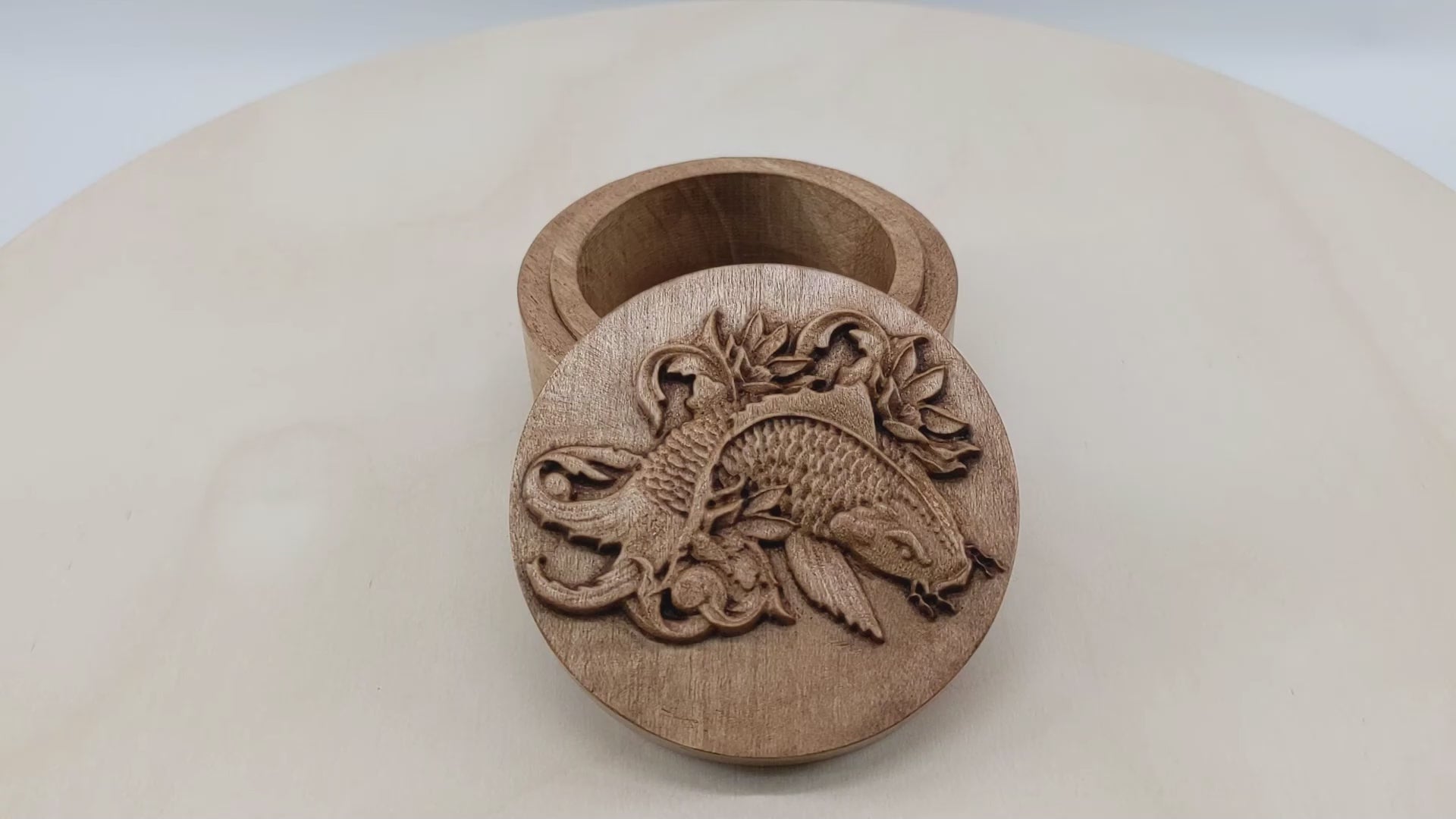 Round wooden box 3D carved with a koi fish surrounded by leaves and filigree designs. It has flowing whiskers, a sharp back fin and intricately carved scales down to its wide tail. Made from hard maple wood stained brown against a white background rotating on a table.