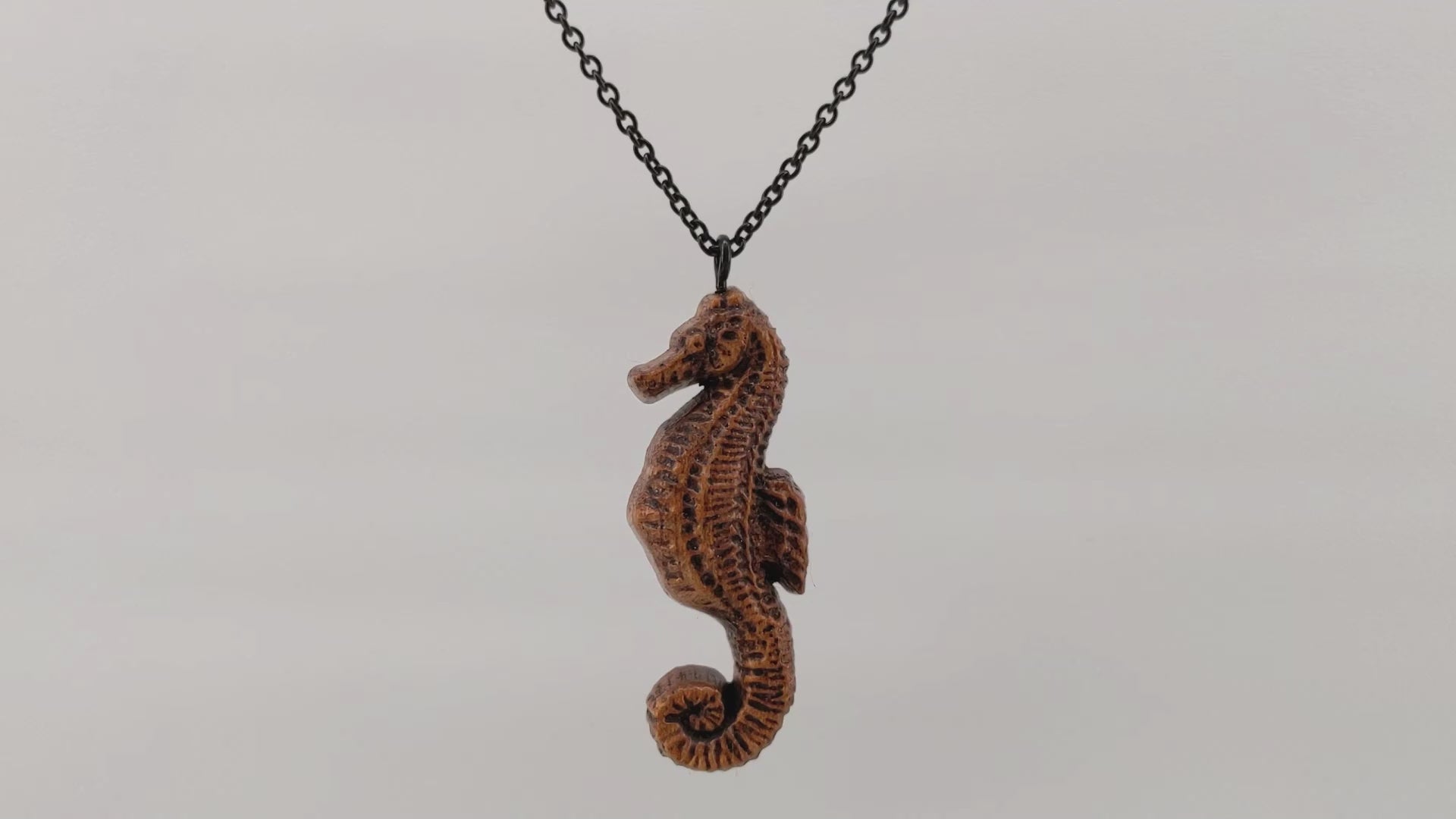 Wooden pendant necklace carved in the shape of sea horse. Made from hard maple wood. Hanging and rotating from a black stainless steel chain against a white background.