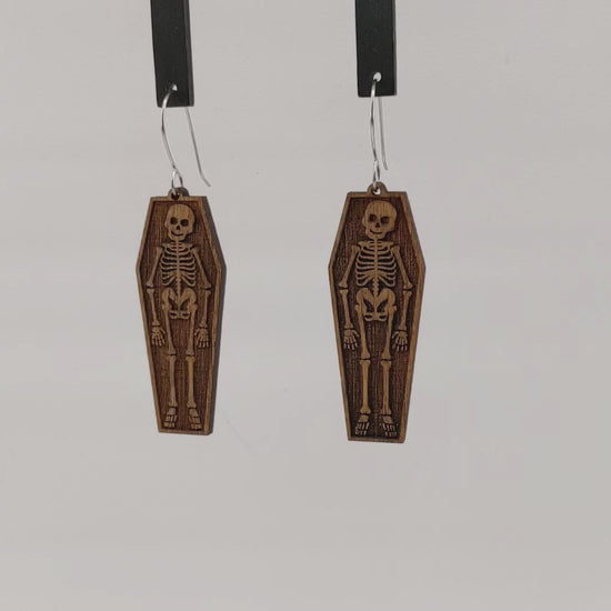 Pair of wooden earrings with silver stainless steel hooks. They are pale, natural finished, engraved skeletons smiling and laying in coffins. Made from birch wood hanging and rotating against white background.