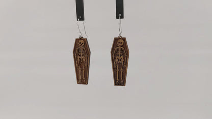 Pair of wooden earrings with silver stainless steel hooks. They are pale, natural finished, engraved skeletons smiling and laying in coffins. Made from birch wood hanging and rotating against white background.