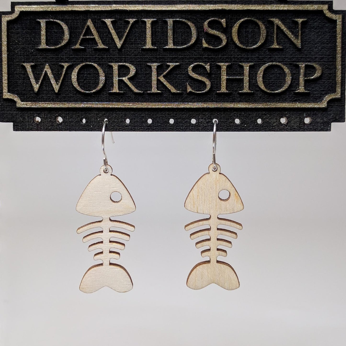 Pair of wooden earrings with silver stainless steel hooks. They are pale natural finished, whimsical simple design,  fish bones. Made from birch wood hanging from a model Davidson Workshop sign against a white background. (Close up View)