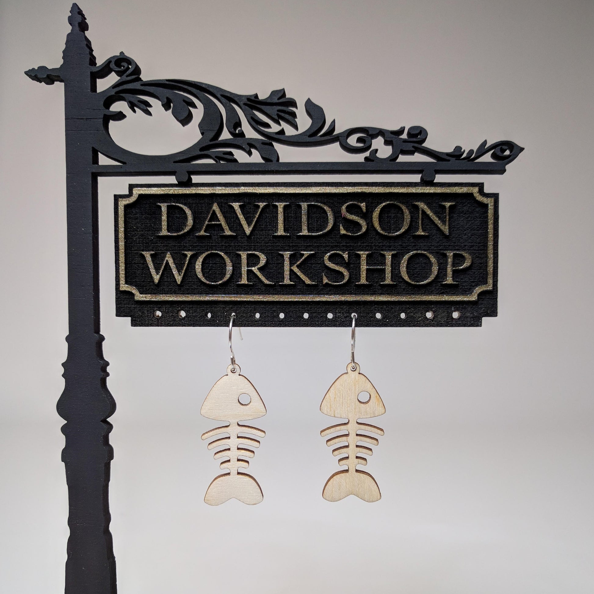 Pair of wooden earrings with silver stainless steel hooks. They are pale natural finished, whimsical simple design,  fish bones. Made from birch wood hanging from a model Davidson Workshop sign against a white background. (Close up View)