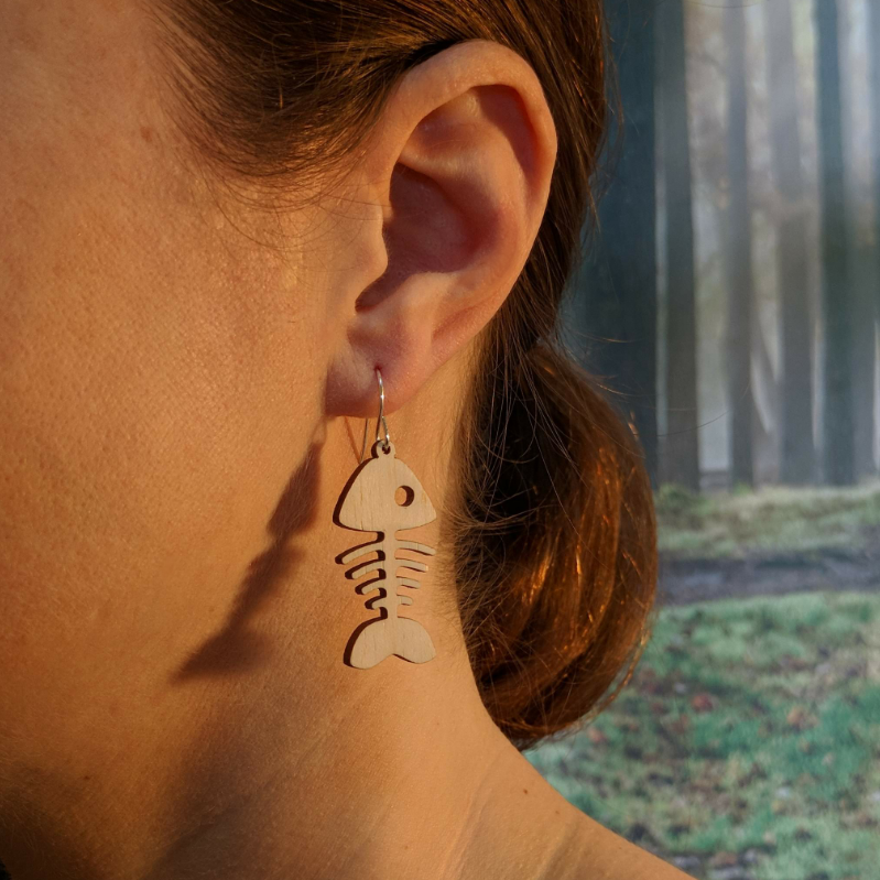 Pair of wooden earrings with silver stainless steel hooks. They are pale natural finished, whimsical simple design, fish bones. Made from birch wood, being modeled by a women in front of a nature scene.