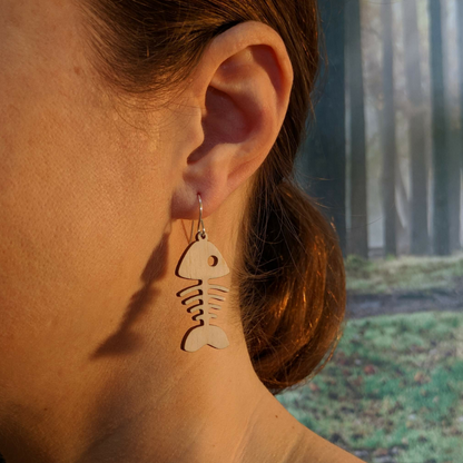 Pair of wooden earrings with silver stainless steel hooks. They are pale natural finished, whimsical simple design, fish bones. Made from birch wood, being modeled by a women in front of a nature scene.