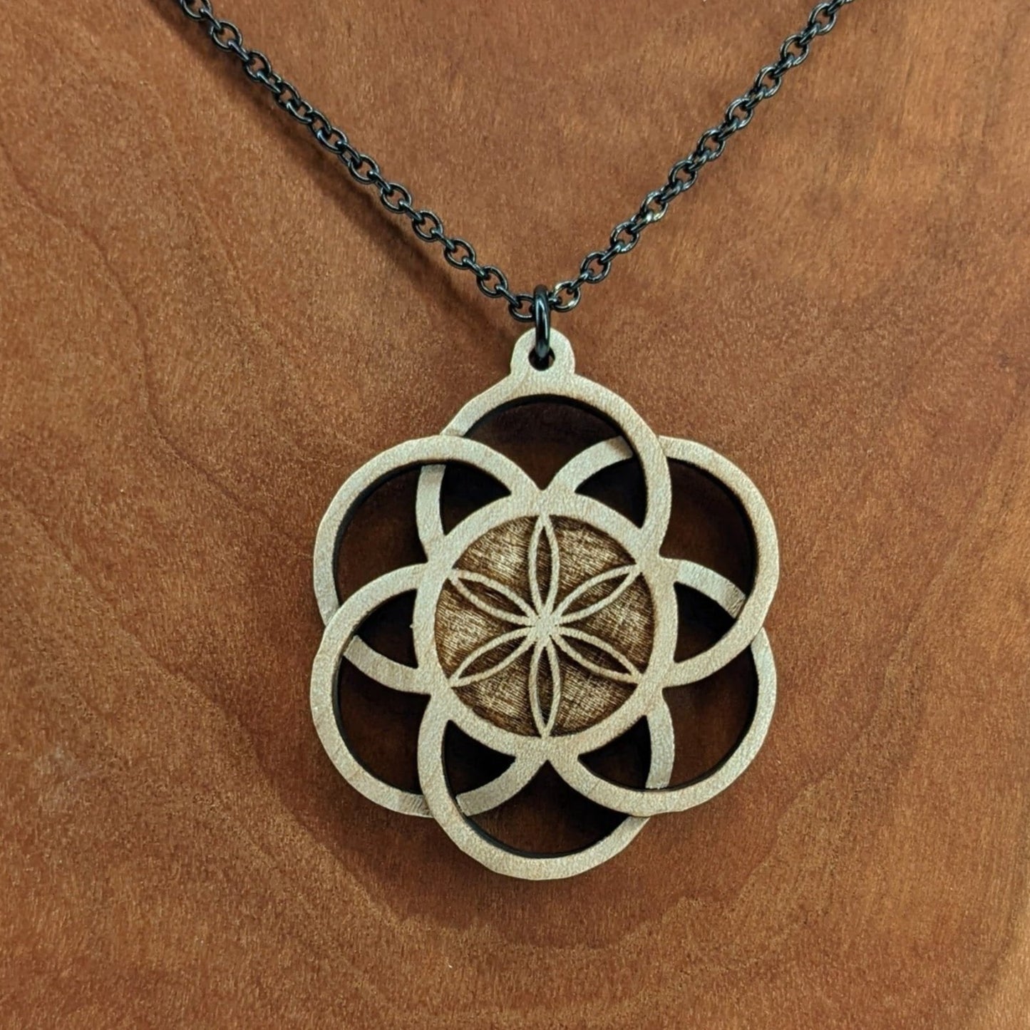 Wooden necklace pendant inspired by a flower of life including 6 interlinking circles creating a flower in the center. Carved from hard maple wood. Hanging from a black stainless steel chain against a cherry wood background.