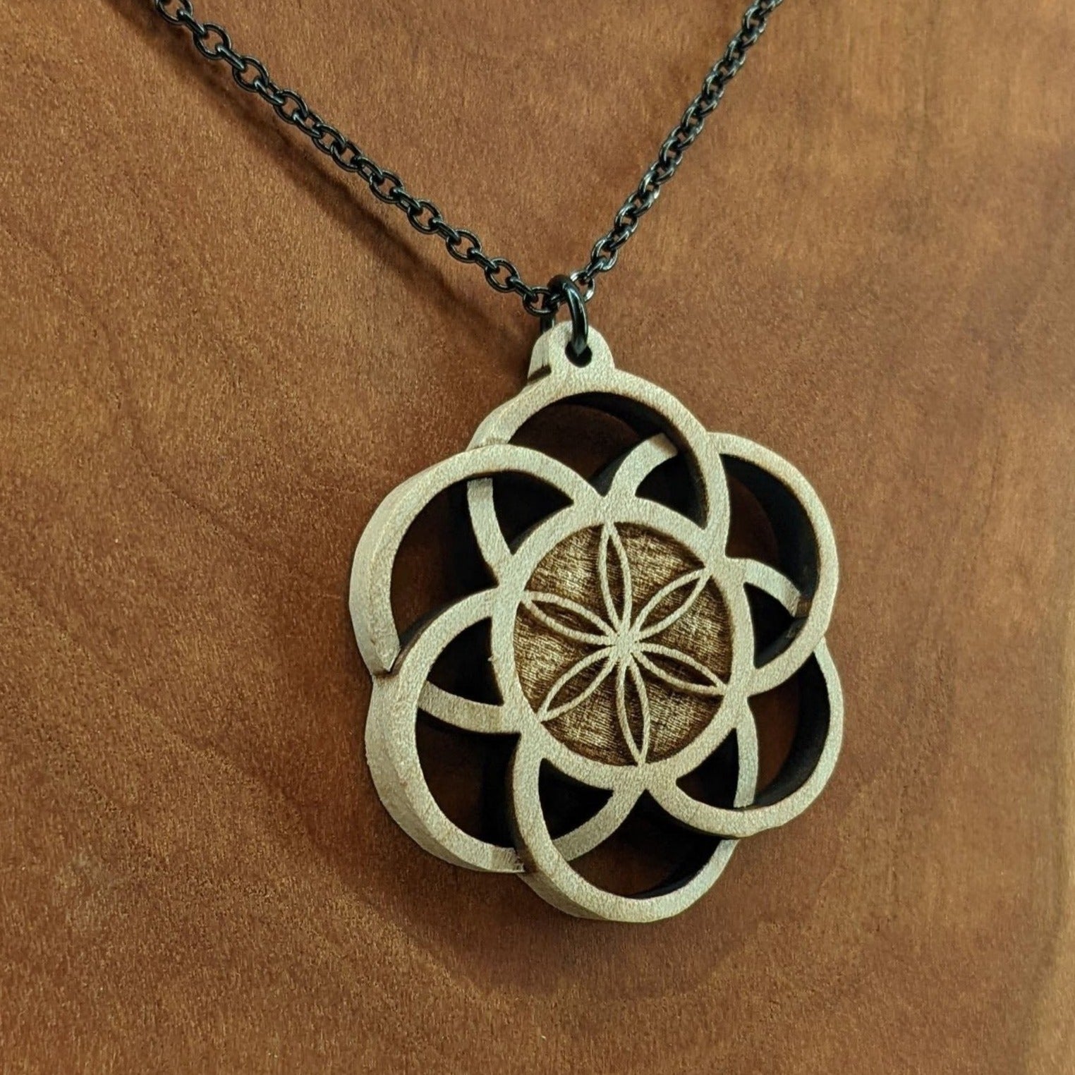 Wooden necklace pendant inspired by a flower of life including 6 interlinking circles creating a flower in the center. Carved from hard maple wood. Hanging from a black stainless steel chain against a cherry wood background.