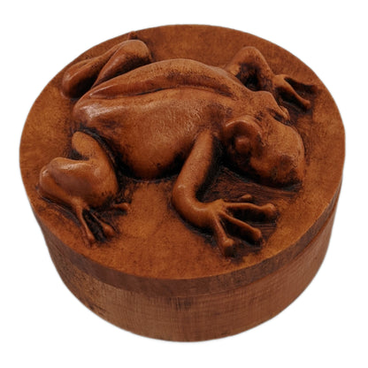 Round wooden box 3D carved with a tree frog in a clinging position. It has smooth skin and long skinny limbs and hands with bulbous fingertips Made from hard maple wood with a brown stain against a white background.