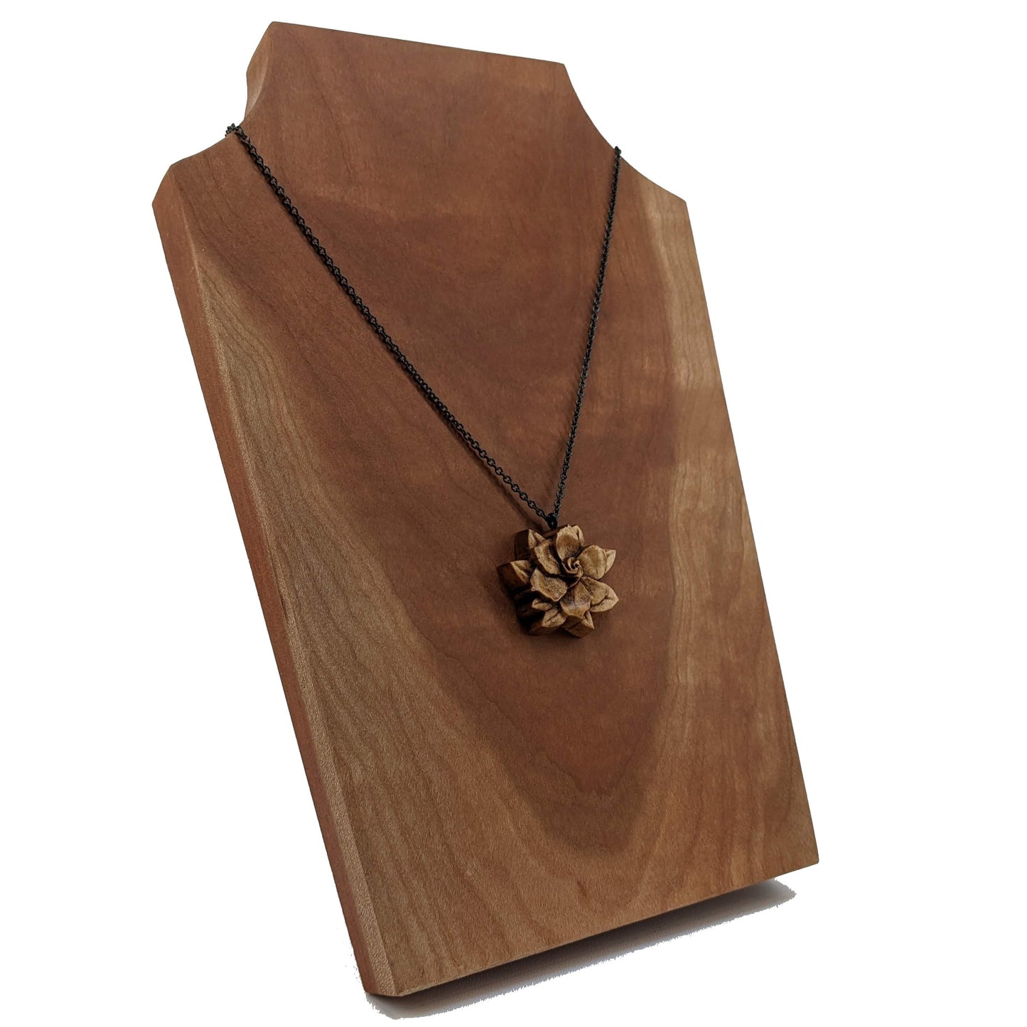 Wooden flower necklace pendant carved in the shape of a gardenia against. Made from hard maple and hanging from a black stainless steel chain against a cherry wood background.
