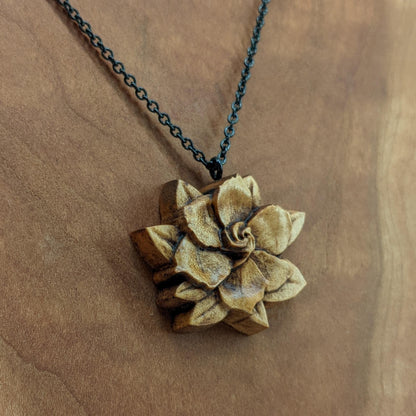 Wooden flower necklace pendant carved in the shape of a gardenia against. Made from hard maple and hanging from a black stainless steel chain against a cherry wood background.