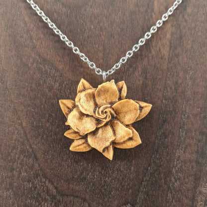 Wooden flower necklace pendant carved in the shape of a gardenia against. Made from hard maple and hanging from a silver stainless steel chain against a dark walnut wood background.