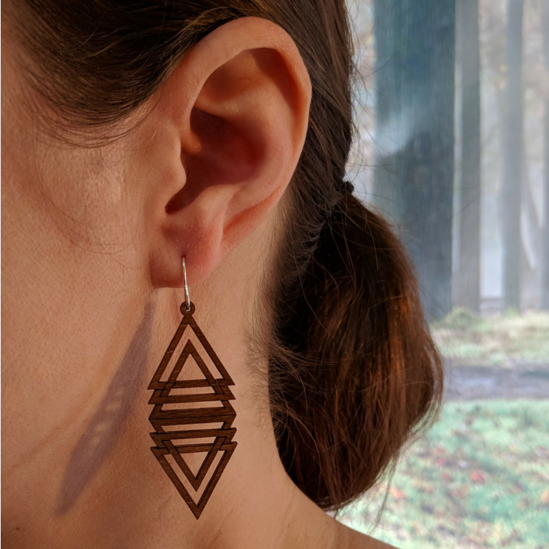 Pair of wooden earrings with silver stainless steel hooks. They are multiple brown triangles overlapping and mirrored to make a diamond shape. Made from birch wood, being modeled by a women in front of a nature scene.