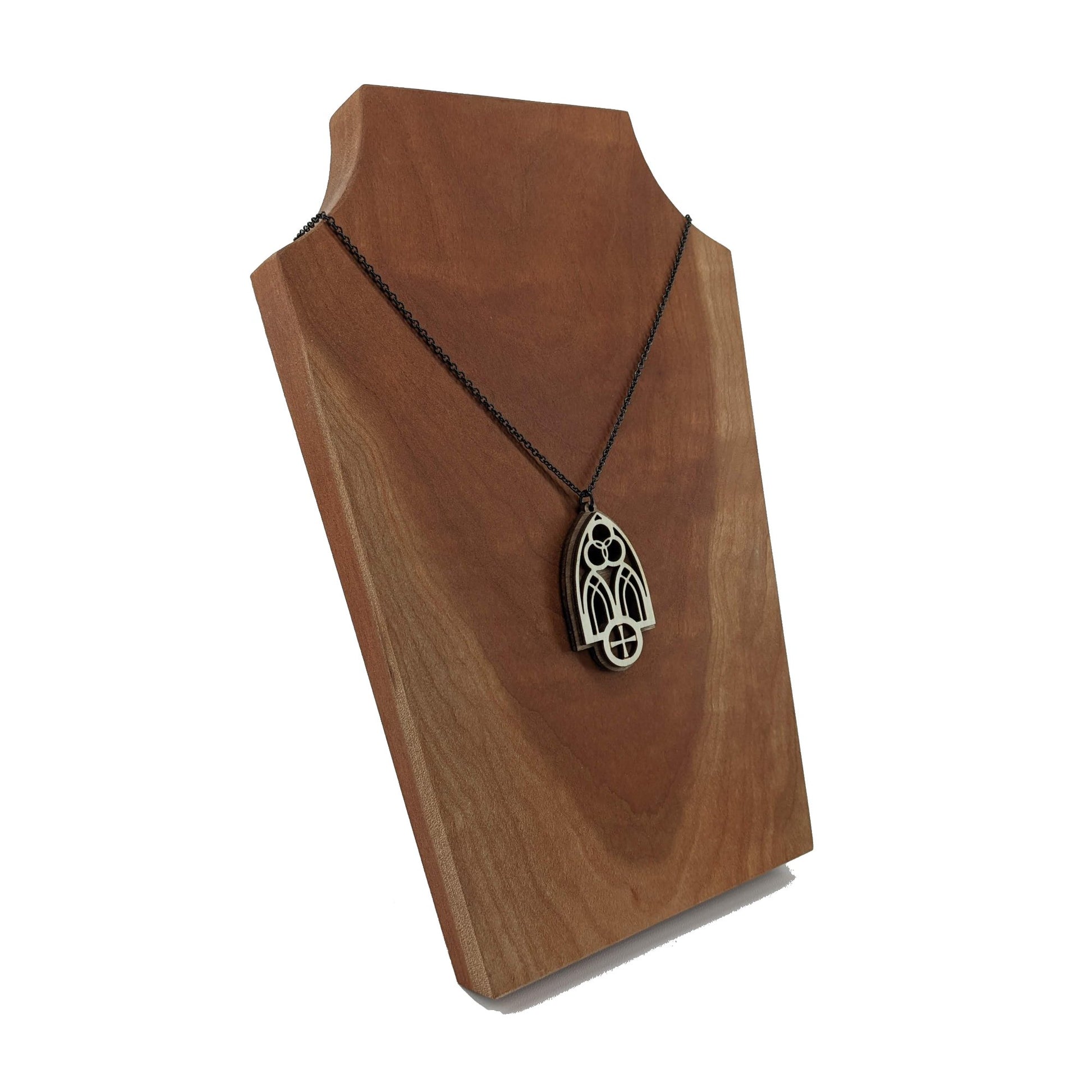 Wooden necklace pendant inspired by a gothic church window. includes a trinity symbol above and a cross below. Made from two layers, dark and light birch wood. Hanging from a black stainless steel chain against a cherry wood display.