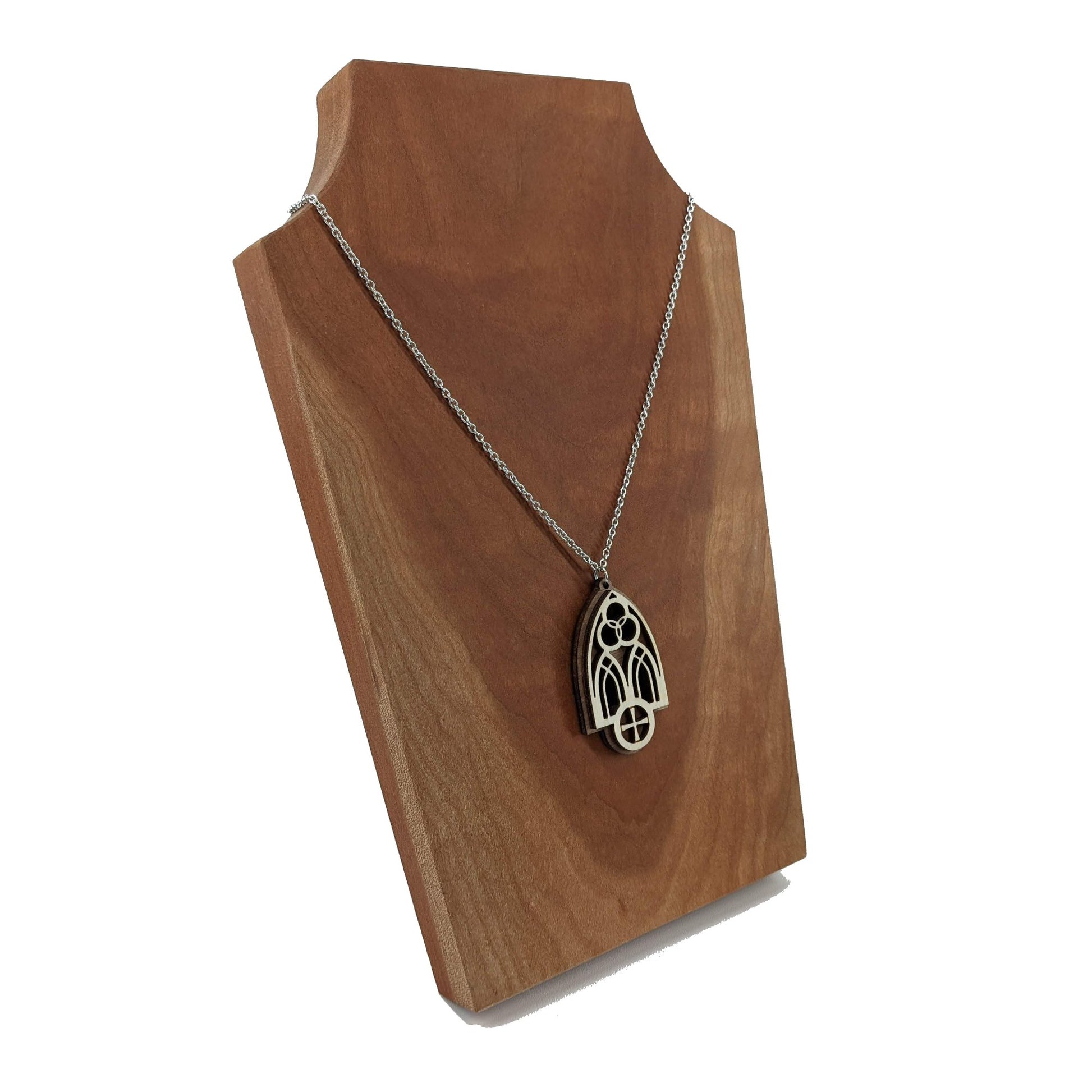 Wooden necklace pendant inspired by a gothic church window. includes a trinity symbol above and a cross below. Made from two layers, dark and light birch wood. Hanging from a silver stainless steel chain against a cherry wood display.