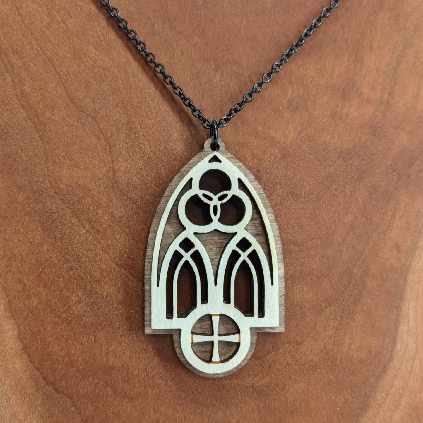 Wooden necklace pendant inspired by a gothic church window. includes a trinity symbol above and a cross below. Made from two layers, dark and light birch wood. Hanging from a black stainless steel chain against a cherry wood background.