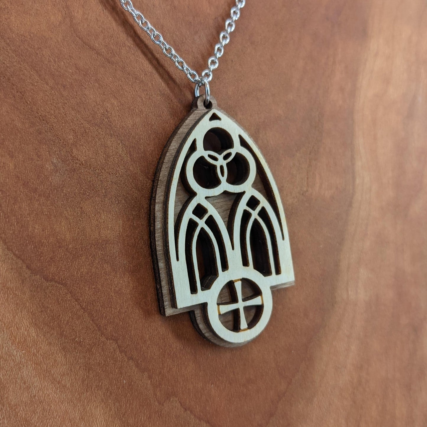 Wooden necklace pendant inspired by a gothic church window. includes a trinity symbol above and a cross below. Made from two layers, dark and light birch wood. Hanging from a silver stainless steel chain against a cherry wood background.