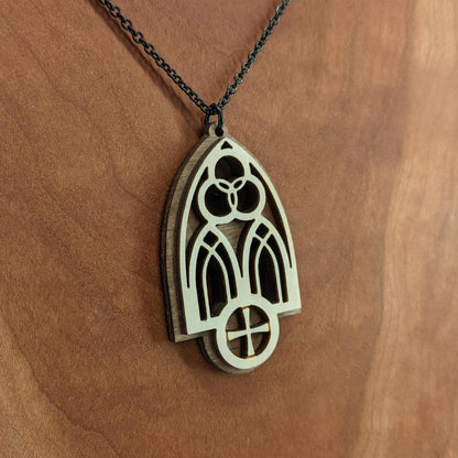 Wooden necklace pendant inspired by a gothic church window. includes a trinity symbol above and a cross below. Made from two layers, dark and light birch wood. Hanging from a black stainless steel chain against a cherry wood background.