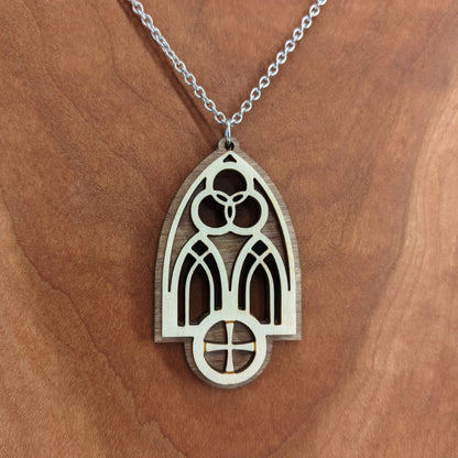 Wooden necklace pendant inspired by a gothic church window. includes a trinity symbol above and a cross below. Made from two layers, dark and light birch wood. Hanging from a silver stainless steel chain against a cherry wood background.