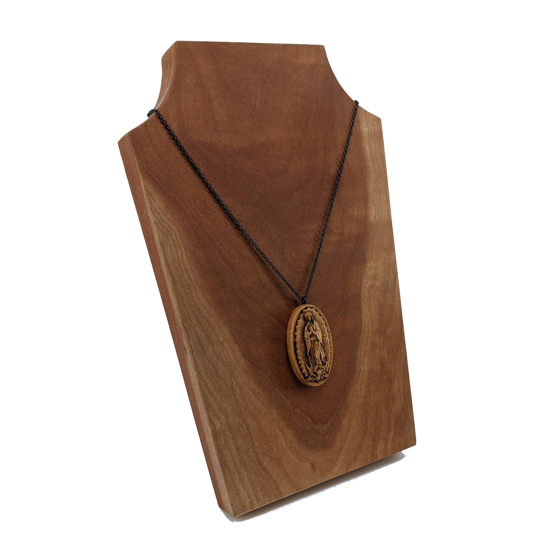 Oval wooden necklace pendant carved in the shape of Guadalupe. Made from hard maple and hanging from a black stainless steel chain against a cherry wood  background.