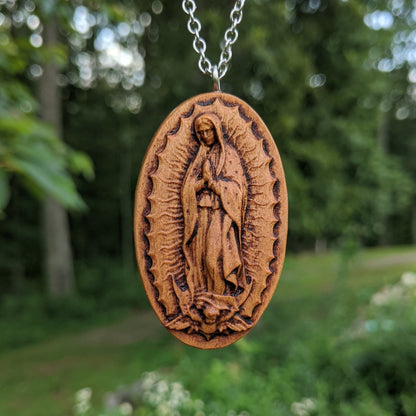 Oval wooden necklace pendant carved in the shape of Guadalupe. Made from hard maple and hanging from a silver stainless steel chain against a forest background.