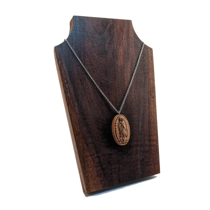 Oval wooden necklace pendant carved in the shape of Guadalupe. Made from hard maple and hanging from a silver stainless steel chain against a dark walnut background.