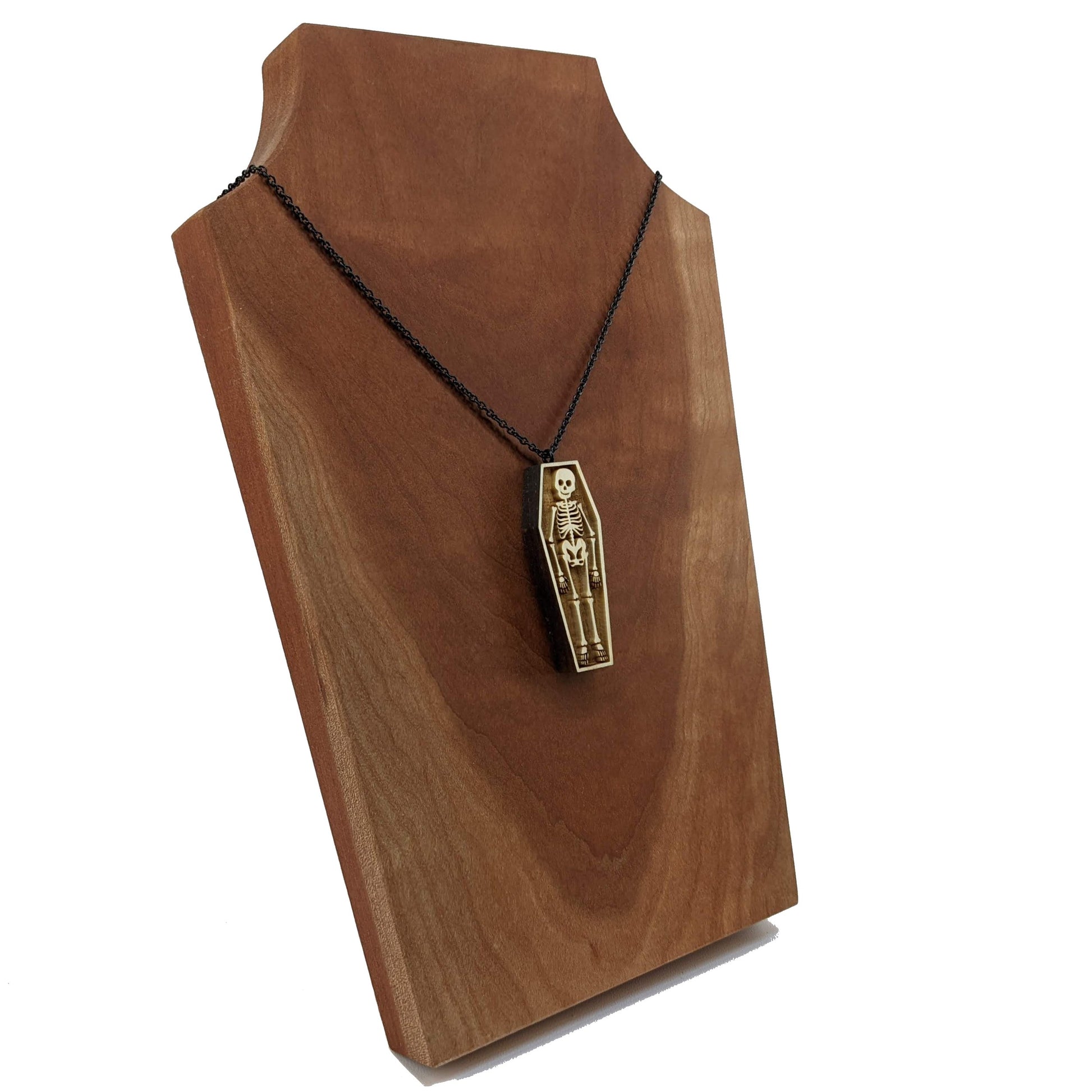 Wooden necklace pendant created in the shape a skeleton lying in a coffin. Hanging from a black stainless steel chain against a cherry wood background.