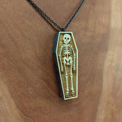 Wooden necklace pendant created in the shape a skeleton lying in a coffin. Hanging from a black stainless steel chain against a cherry wood background.