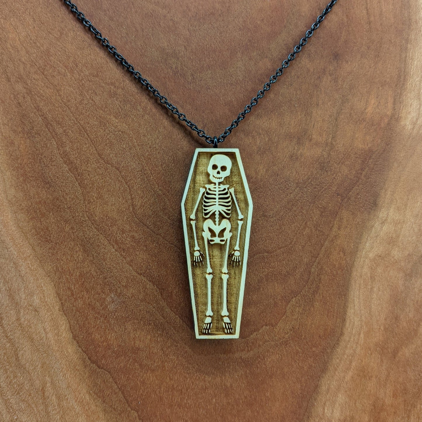 Wooden necklace pendant created in the shape a skeleton lying in a coffin. Hanging from a black stainless steel chain against a cherry wood background.