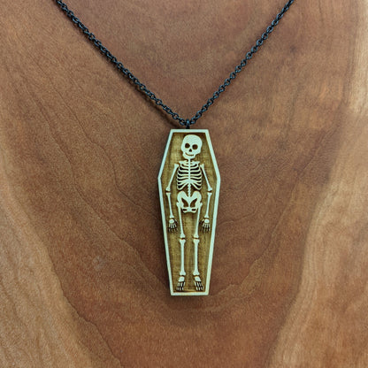 Wooden necklace pendant created in the shape a skeleton lying in a coffin. Hanging from a black stainless steel chain against a cherry wood background.