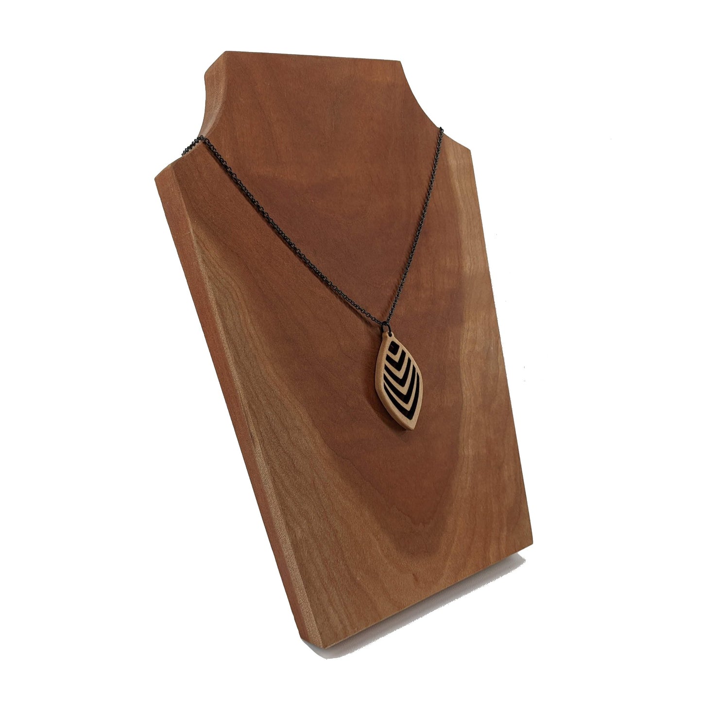 Wooden necklace pendant cut into a teardrop shape with a chevron pattern cut throughout it. Made from hard maple and hanging from a black stainless steel chain against a cherry wood background.