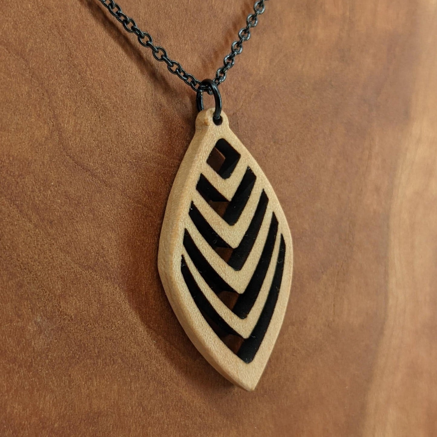 Wooden necklace pendant cut into a teardrop shape with a chevron pattern cut throughout it. Made from hard maple and hanging from a black stainless steel chain against a cherry wood background.