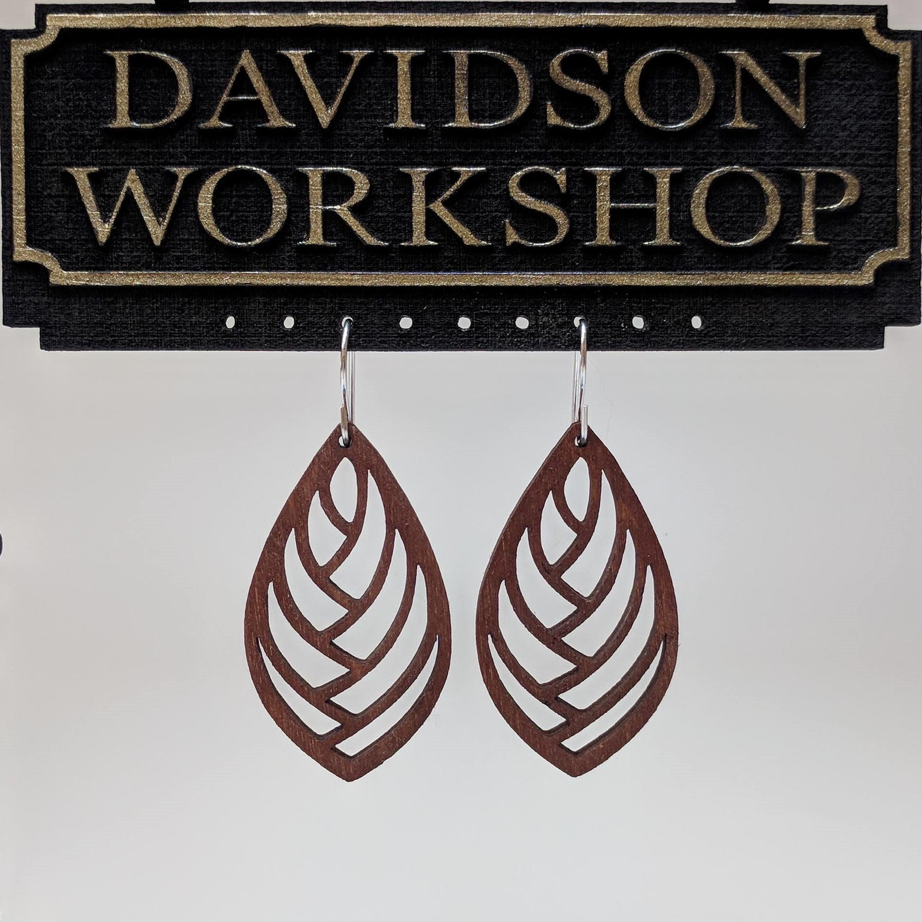 Pair of wooden earrings with silver stainless steel hooks. They are brown almond shaped cutouts with a herringbone pattern within. Made from birch wood hanging from a model Davidson Workshop sign against a white background. (Close up View