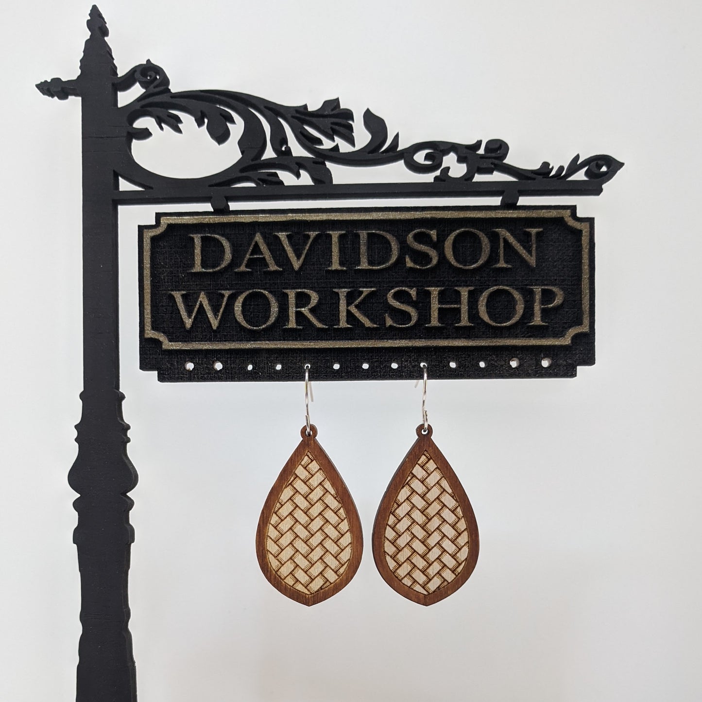 Pair of wooden earrings with silver stainless steel hooks. They are brown almond shaped with an engraved herringbone weave pattern within. Made from birch wood hanging from a model Davidson Workshop sign against a white background.