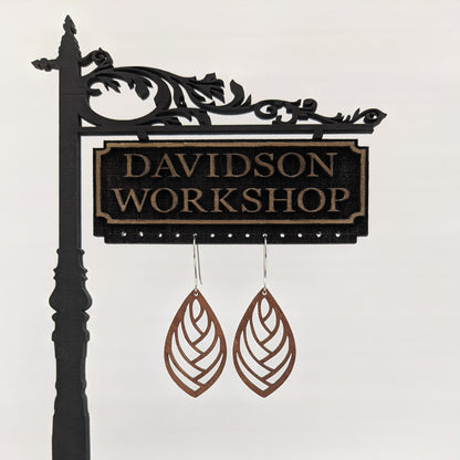 Pair of wooden earrings with silver stainless steel hooks. They are brown almond shaped with a herringbone style cutout within. Made from birch wood hanging from a model Davidson Workshop sign against a white background. (Close up View)