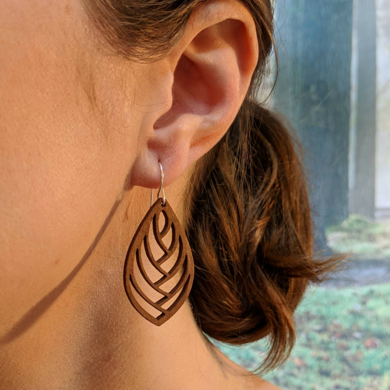 Pair of wooden earrings with silver stainless steel hooks. They are brown almond shaped with a herringbone style cutout within. Made from birch wood, being modeled by a women in front of a nature scene.