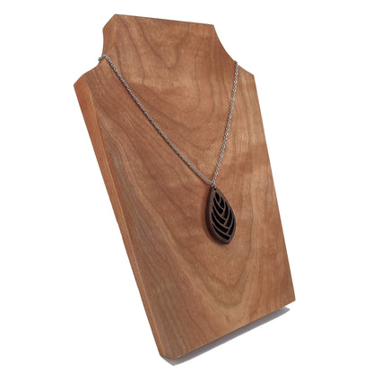 Wooden necklace pendant cut into a teardrop shape with a herring bone pattern cut throughout it. Made from walnut and hanging from a silver stainless steel chain against a cherry wood background.