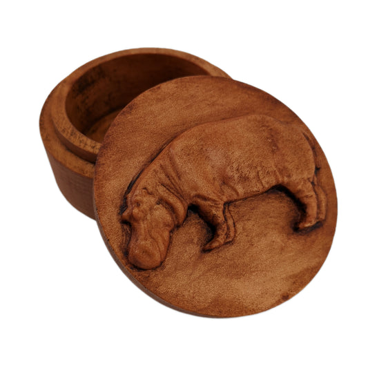 Round wooden box 3D carved with a hippopotamus standing broadside to the left. Its head is hanging downward as if drinking from a stream. Made from hard maple wood stained brown against a white background.