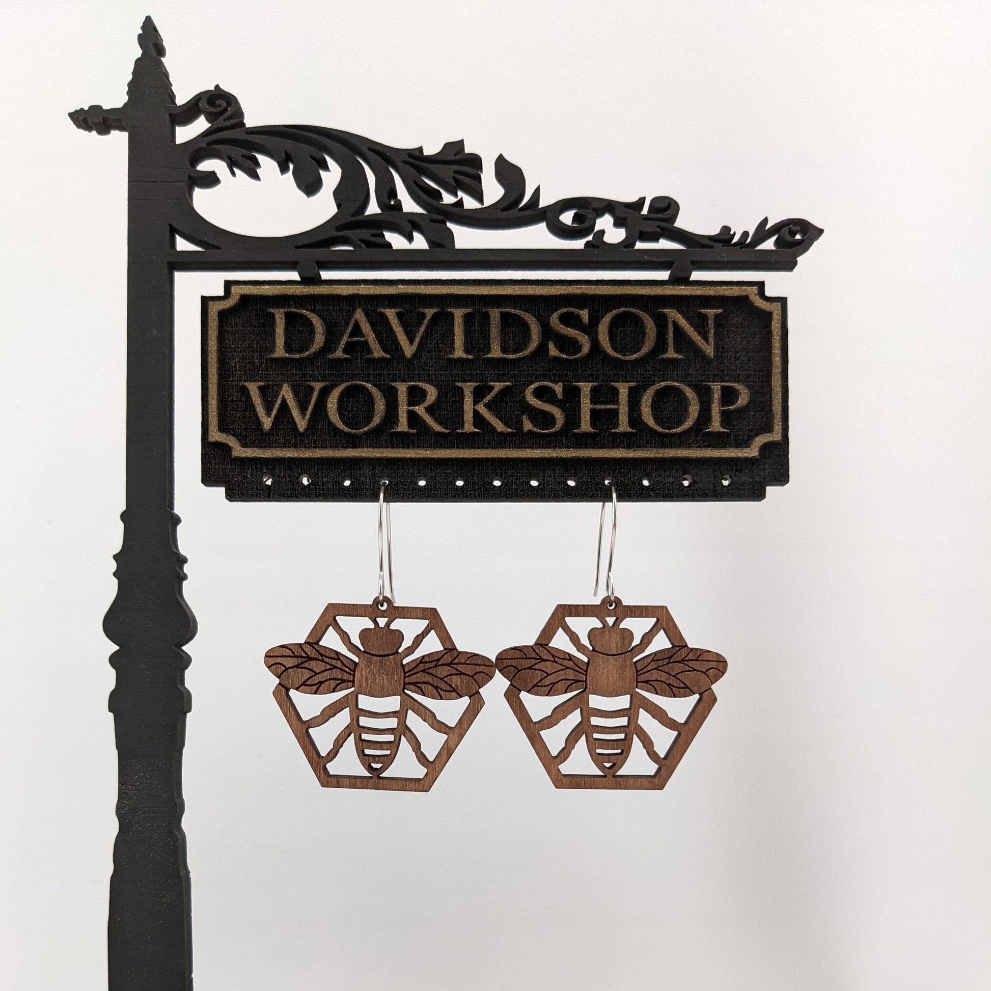 Pair of wooden earrings with silver stainless steel hooks. They are stained brown honeybees with its arms and wings spread on a honeycomb. Made from birch wood hanging from a model Davidson Workshop sign against a white background.