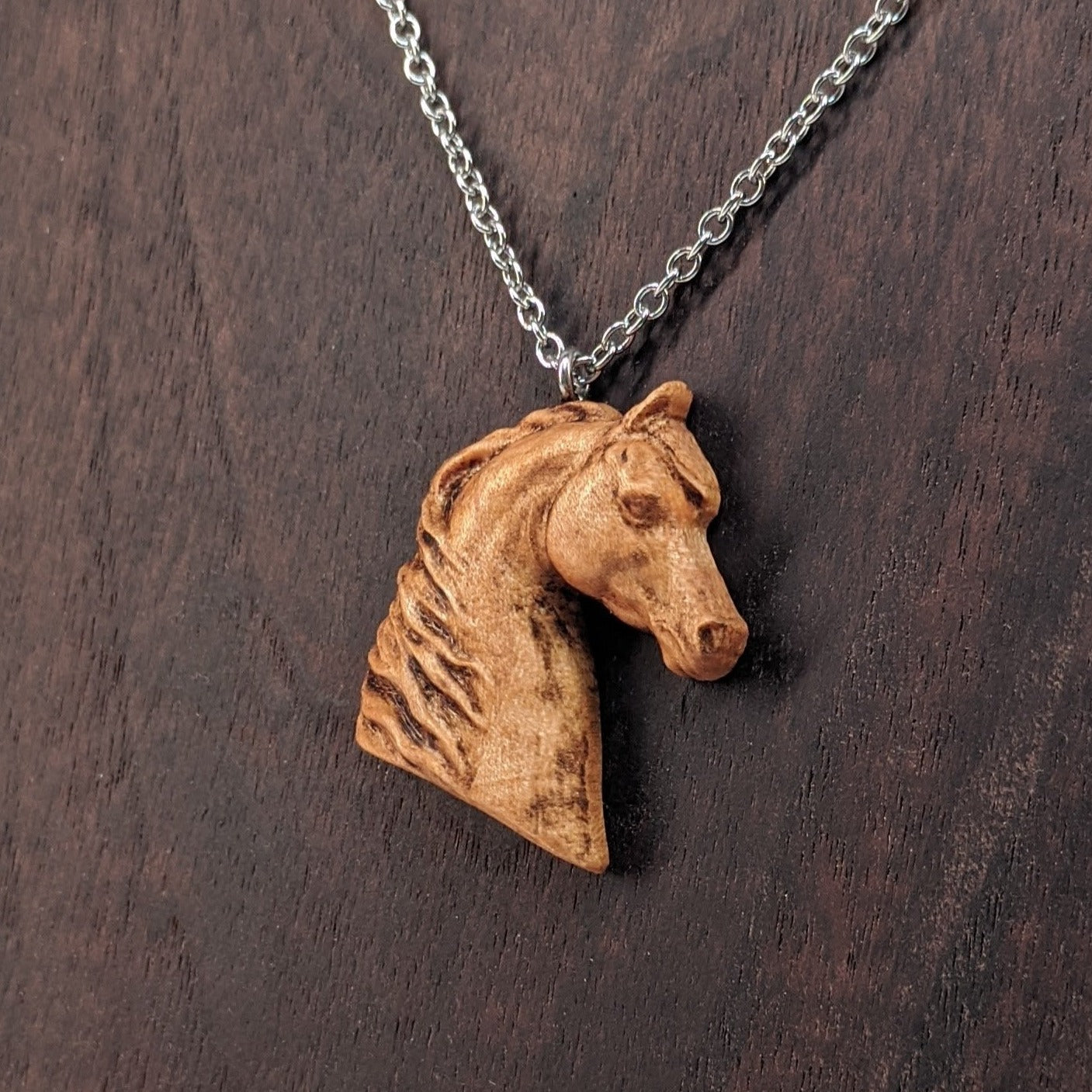 Wooden pendant realistically carved in the shape of a horse. Made form hard maple and hung from a silver stainless steel chain against a dark walnut background.