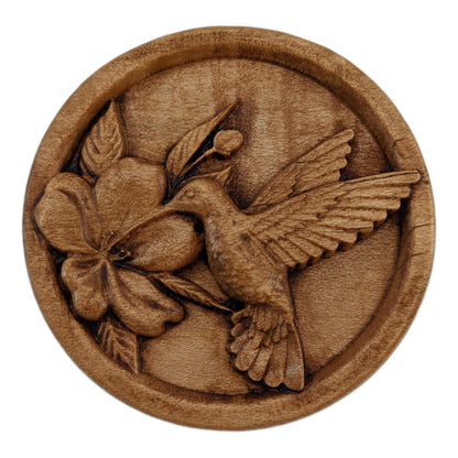 Round wooden box 3D carved with a recessed hummingbird hovering and drinking nectar from a flower. It is facing to the left with its delicate wings aflutter as it leans in to drink. Made from hard maple wood stained brown against a white background.