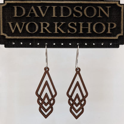 Pair of wooden earrings with silver stainless steel hooks. They are brown elongated diamonds, overlapping each other to form a woven pattern. Made from birch wood hanging from a model Davidson Workshop sign against a white background (Close up View)