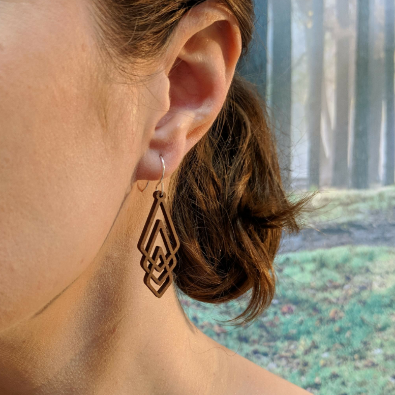 Pair of wooden earrings with silver stainless steel hooks. They are 3 brown diamond shapes interlocking to make an elongated shard shape. Made from birch wood, being modeled by a women in front of a nature scene.