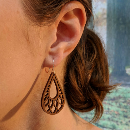 Pair of wooden earrings with silver stainless steel hooks. They are brown teardrop shaped with a woven pattern within and a tear in the center. Made from birch wood, being modeled by a women in front of a nature scene.