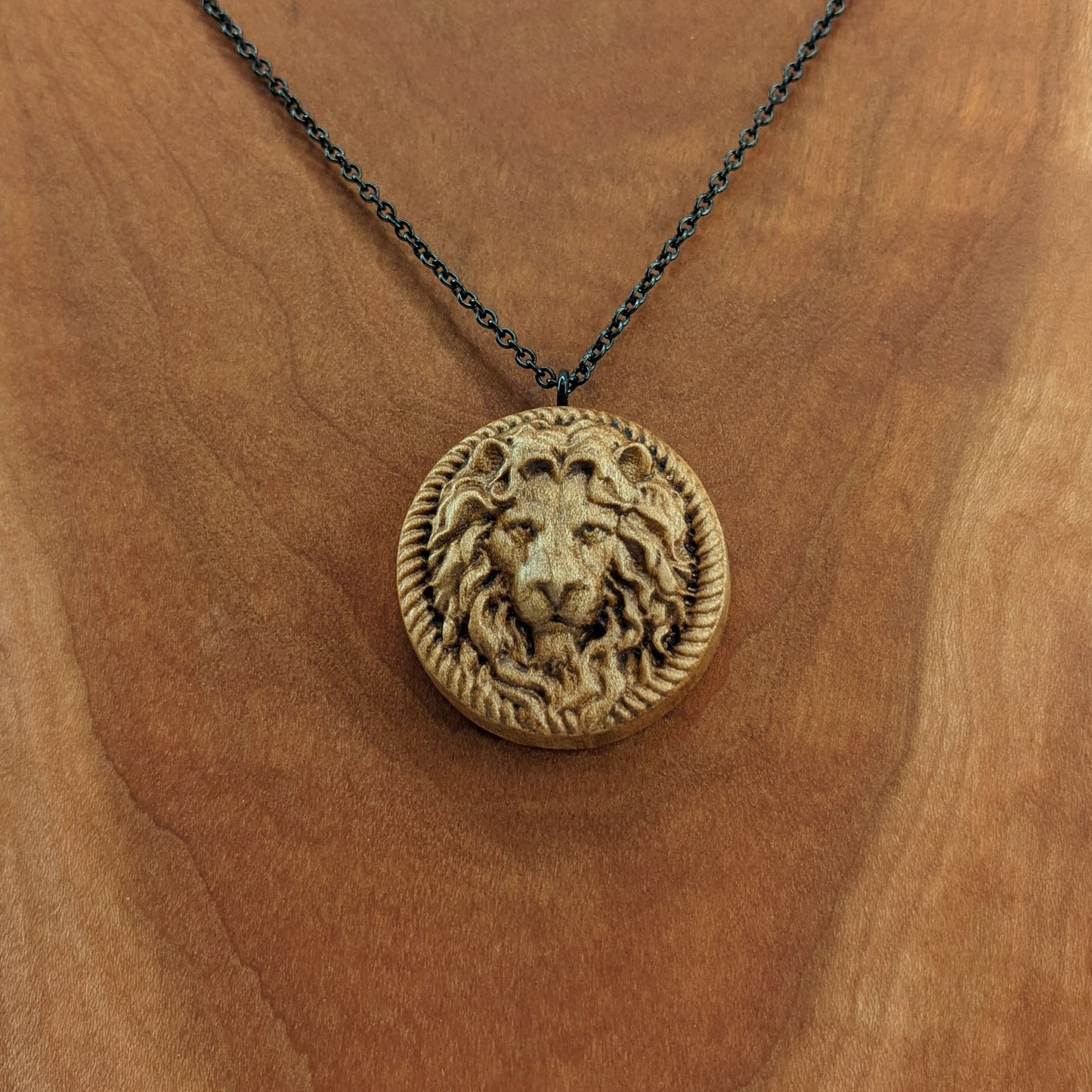 Wooden necklace pendant carved in the shape of a stern lion face surrounded by a rope border. Hanging from a black stainless steel chain against a cherry wood background.